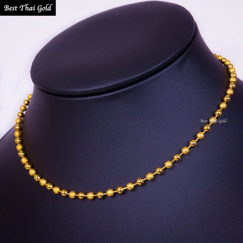 Pu&ograve; includere: Una collana a catena con palline dorate composta da una serie di piccole perle sferiche testurizzate collegate tra loro. La collana &egrave; esposta su un supporto per collo scuro. Il testo "Best Thai Gold" &egrave; visibile in alto a sinistra e a destra.