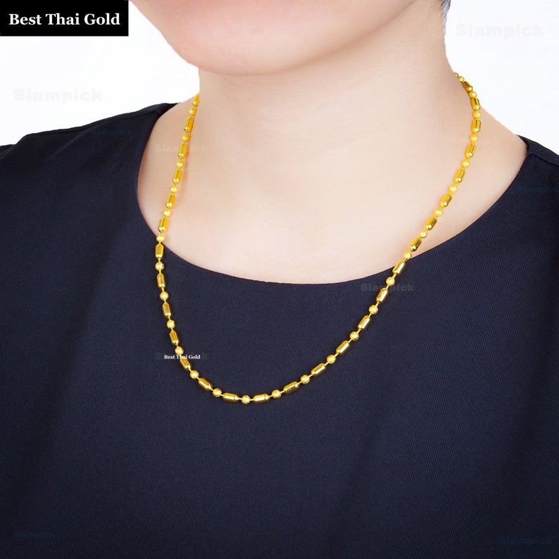May include: A yellow gold chain necklace featuring an alternating pattern of small spherical beads and elongated cylindrical links. The necklace is displayed against a plain black garment. The text Best Thai Gold is visible in the top corner.