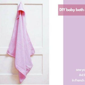 Puede incluir: Una capa de baño para bebé de color rosa claro colgada de una puerta blanca. La capa tiene una capucha y está hecha de tela suave. El texto a la derecha dice "DIY baby bath cape" y "sew your own!" en francés e inglés.