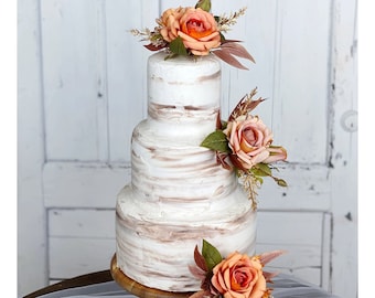 May include: A three-tiered wedding cake with a white buttercream frosting and a rustic woodgrain design. The cake is decorated with peach-colored roses and greenery.