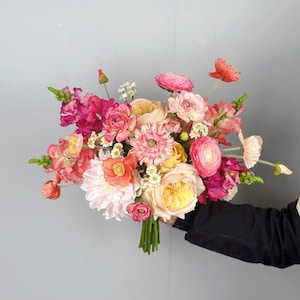 May include: A vibrant bouquet of mixed flowers in shades of pink, peach, and yellow. The arrangement includes roses, dahlias, and other blooms, held together with green stems. The flowers are set against a gray background.
