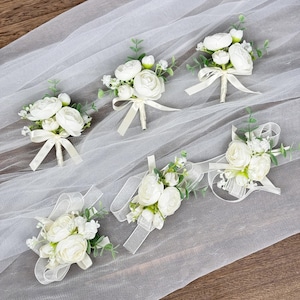 May include: Six white artificial flower buttonholes with green leaves and white ribbon accents on a white tulle background.