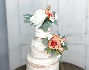 May include: A three-tiered white wedding cake with a rustic woodgrain finish, decorated with peach and white flowers and greenery. The cake is displayed on a black pedestal stand.
