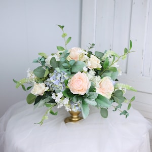 Könnte beinhalten: Eine Tischdekoration mit künstlichen Blumen in einer goldenen Vase. Die Dekoration besteht aus Pfirsich- und weißen Rosen, blauen Hortensien und Grün.