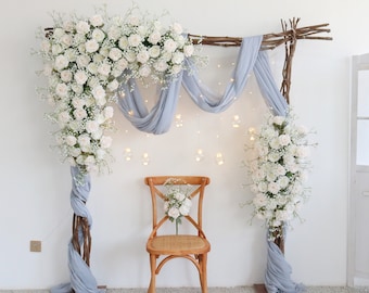 May include: A white wooden archway decorated with white flowers and a light blue sheer fabric. A wooden chair sits in front of the archway.