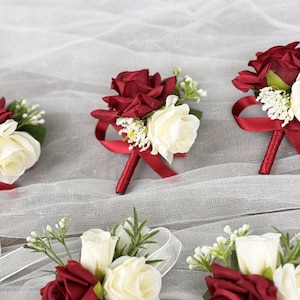 May include: Five burgundy and white rose boutonnieres with red ribbon accents. The boutonnieres are arranged on a white tulle fabric.
