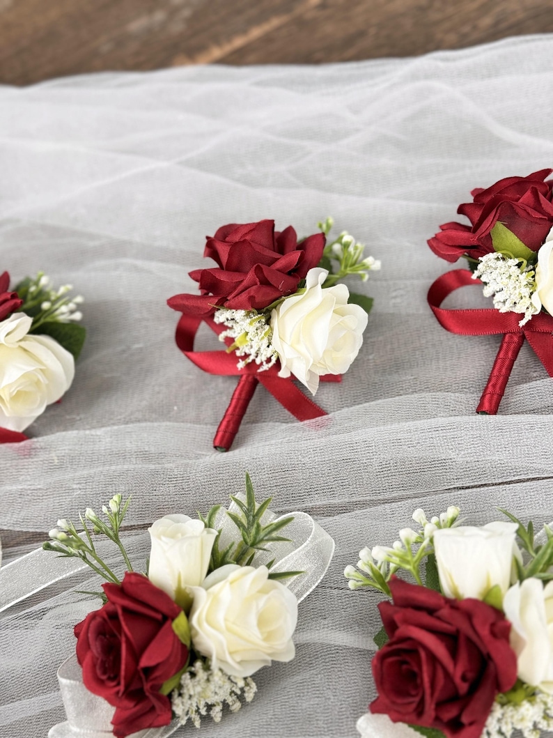 May include: Five burgundy and white rose boutonnieres with red ribbon accents. The boutonnieres are arranged on a white tulle fabric.