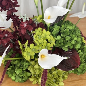 May include: A floral arrangement featuring white calla lilies with yellow centers, deep burgundy flowers, and clusters of bright green hydrangea-like blooms. The composition includes long green stems and dark red, glossy, heart-shaped leaves.