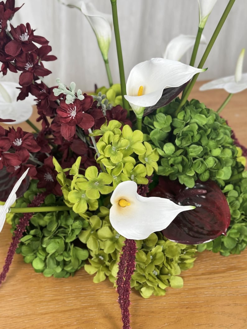 May include: A floral arrangement featuring white calla lilies with yellow centers, deep burgundy flowers, and clusters of bright green hydrangea-like blooms. The composition includes long green stems and dark red, glossy, heart-shaped leaves.