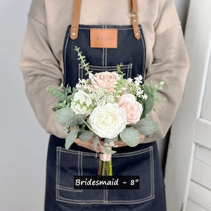 May include: A bridesmaid bouquet featuring white and blush pink roses, greenery, and white baby's breath. The bouquet measures 8 inches in diameter and is tied with a white ribbon. Bridesmaid - 8"