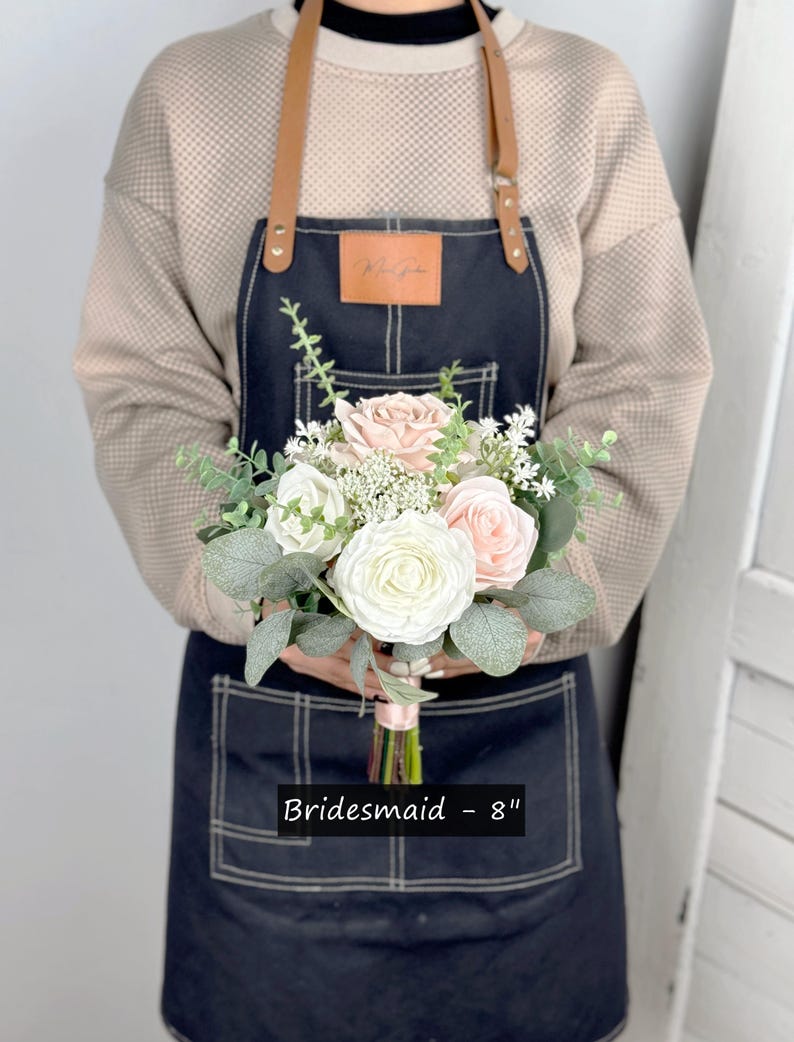 May include: A bridesmaid bouquet featuring white and blush pink roses, greenery, and white baby's breath. The bouquet measures 8 inches in diameter and is tied with a white ribbon. Bridesmaid - 8"
