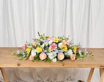 May include: A vibrant floral arrangement featuring a variety of colorful blooms, including pink, yellow, and blue flowers, arranged on a wooden table. The flowers are set against a white backdrop.