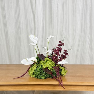 May include: A floral arrangement featuring white anthurium flowers, vibrant green hydrangea clusters, deep burgundy larkspur, and long, thin, dark red amaranth strands. The composition is dense and rounded, showcasing a mix of textures.