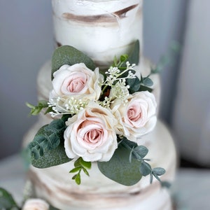 May include: A three-tier wedding cake decorated with blush pink roses and greenery. The cake is a semi-naked cake with a rustic, natural look.