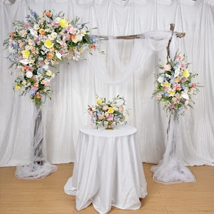 Könnte beinhalten: Hochzeitszeremonie-Arrangement mit floralen Arrangements in Pastellfarben. Die Präsentation umfasst einen Blumenbogen, zwei Blumensäulen und einen kleinen Tisch mit einem Blumengesteck. Weißer Stoff drapiert den Hintergrund und die Säulen.
