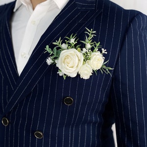 May include: A navy blue pinstripe suit jacket with a white button-down shirt underneath. A white rose boutonniere is pinned to the lapel.