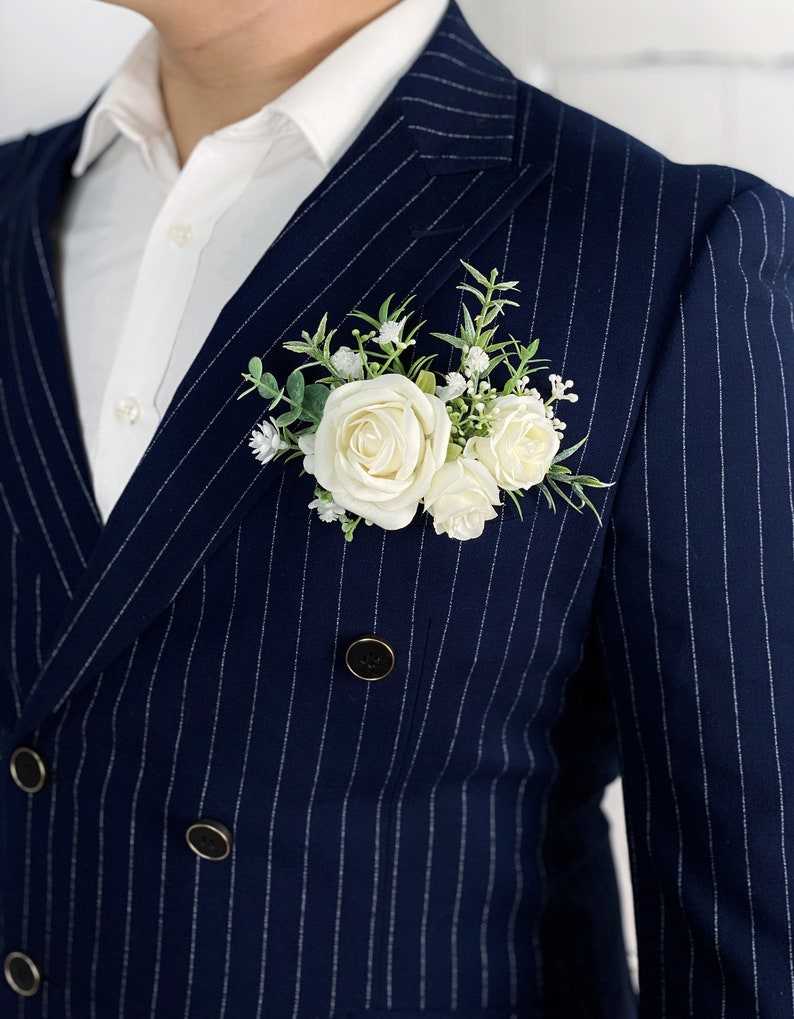 May include: A navy blue pinstripe suit jacket with a white button-down shirt underneath. A white rose boutonniere is pinned to the lapel.