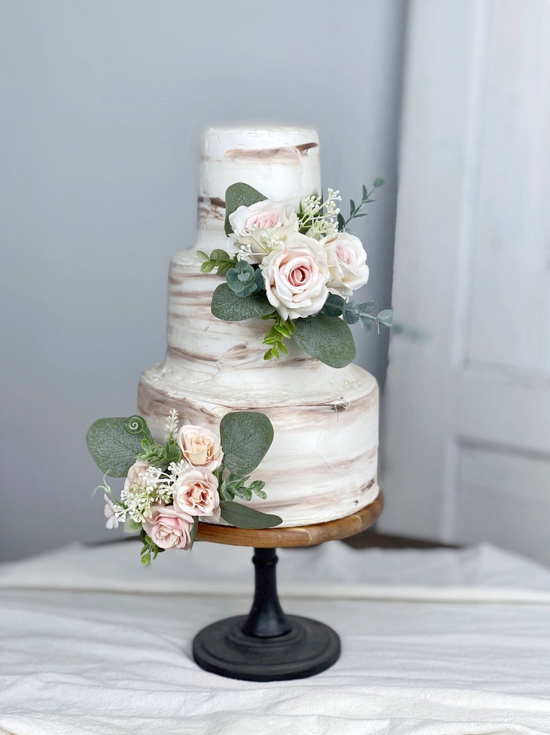 May include: A three-tier wedding cake with white and brown marbled icing. The cake is decorated with pink roses and greenery.
