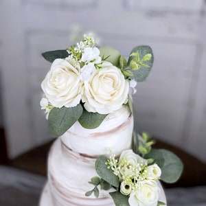 Op de afbeelding: Een drielaagse witte bruidstaart versierd met witte rozen en eucalyptusbladeren.