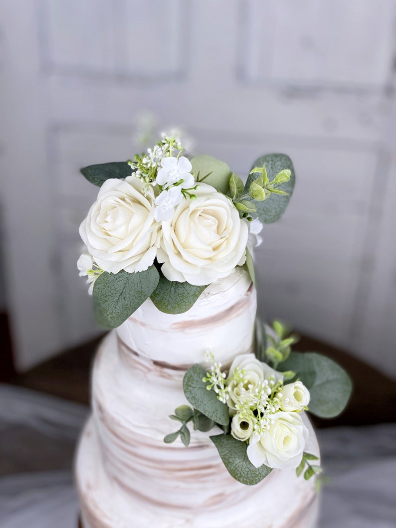 Op de afbeelding: Een drielaagse witte bruidstaart versierd met witte rozen en eucalyptusbladeren.