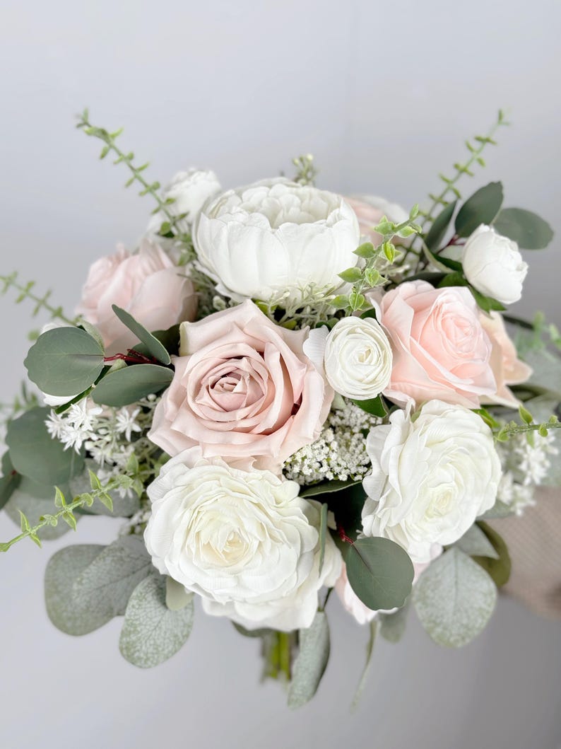 May include: A bridal bouquet featuring white and blush pink roses, white ranunculus, and greenery.