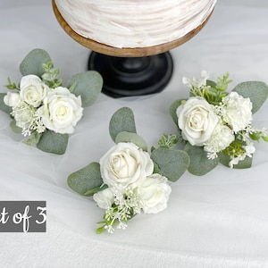 Op de afbeelding: Set van drie witte rozenbloemstukken met groen. Elk bloemstuk heeft een enkele witte roos met kleinere witte bloemen en eucalyptusbladeren. De tekst "Set of 3" is zichtbaar in de afbeelding.