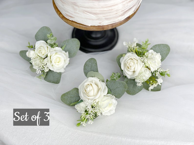 Op de afbeelding: Set van drie witte rozenbloemstukken met groen. Elk bloemstuk heeft een enkele witte roos met kleinere witte bloemen en eucalyptusbladeren. De tekst "Set of 3" is zichtbaar in de afbeelding.