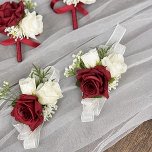 May include: Three red and white rose boutonnieres with red ribbon accents. The boutonnieres are on a white tulle fabric background.