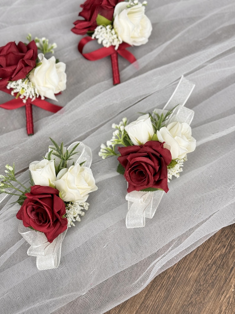 May include: Three red and white rose boutonnieres with red ribbon accents. The boutonnieres are on a white tulle fabric background.