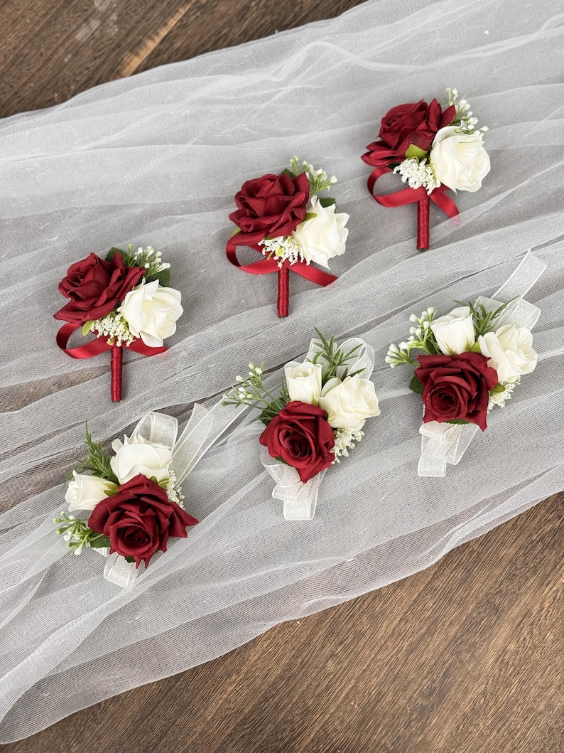 May include: Six burgundy and white artificial rose boutonnieres with red ribbon accents. The boutonnieres are arranged on a white tulle fabric.