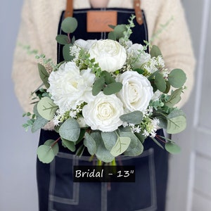 May include: A white bridal bouquet with white roses, peonies, and greenery. The bouquet is 13 inches in diameter and is held by a person wearing a black apron. "Bridal - 13" is written on the apron.