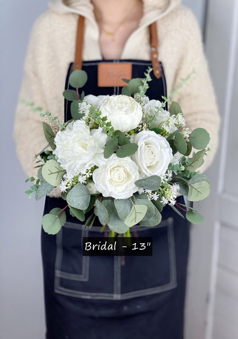 May include: A white bridal bouquet with white roses, peonies, and greenery. The bouquet is 13 inches in diameter and is held by a person wearing a black apron. "Bridal - 13" is written on the apron.