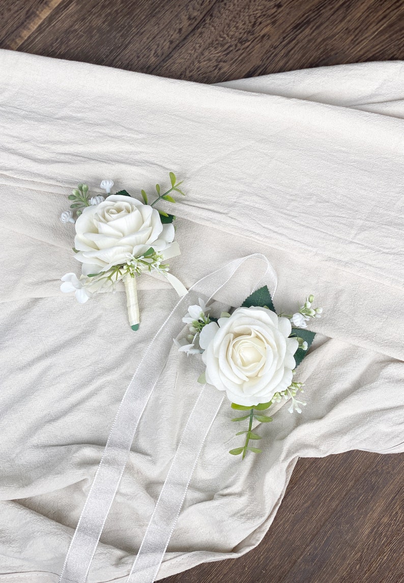 May include: Two white rose boutonnieres with green leaves and white flowers. The boutonnieres are on a white fabric background.