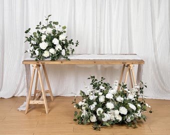 May include: A wooden table with white flowers and greenery arranged on top and below. The table has a light-colored wood top and is supported by wooden sawhorses. The background is a white curtain.