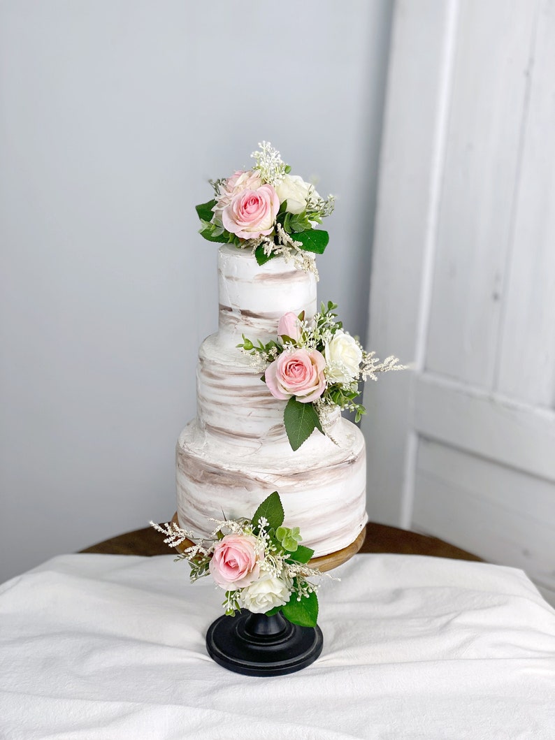 May include: A three-tiered wedding cake with white buttercream frosting and a rustic, marbled design. The cake is decorated with pink and white roses and greenery.