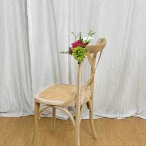 Könnte beinhalten: Ein heller Holzstuhl mit X-förmiger Rückenlehne und einer Sitzfläche aus geflochtenem Rattan. Ein Blumenarrangement mit einer großen lindgrünen Dahlie, leuchtend rosa Blüten und grünem Blattwerk ist mit einem weißen Band an der Lehne befestigt.