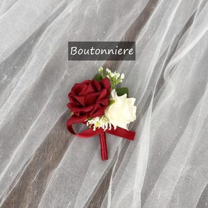 May include: A red and white rose boutonniere with a burgundy ribbon. The boutonniere is on a white tulle fabric.