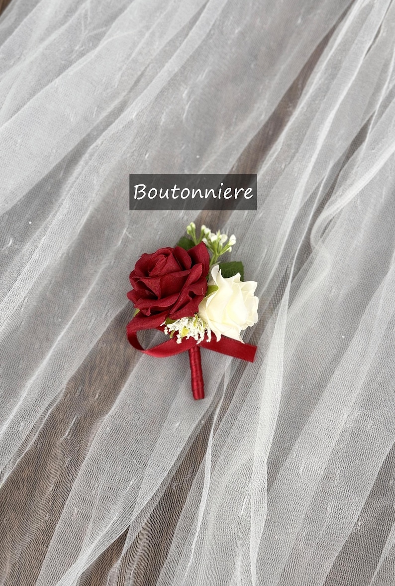 May include: A red and white rose boutonniere with a burgundy ribbon. The boutonniere is on a white tulle fabric.