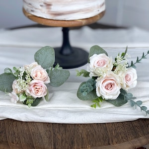 May include: Two small bouquets of blush pink roses and white baby's breath with eucalyptus greenery. The bouquets are arranged on a white tablecloth.