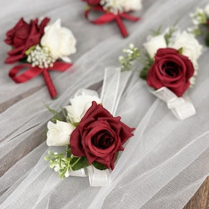 May include: Six burgundy and white artificial flower boutonnieres with red ribbon accents. The boutonnieres are arranged on a white tulle fabric background.