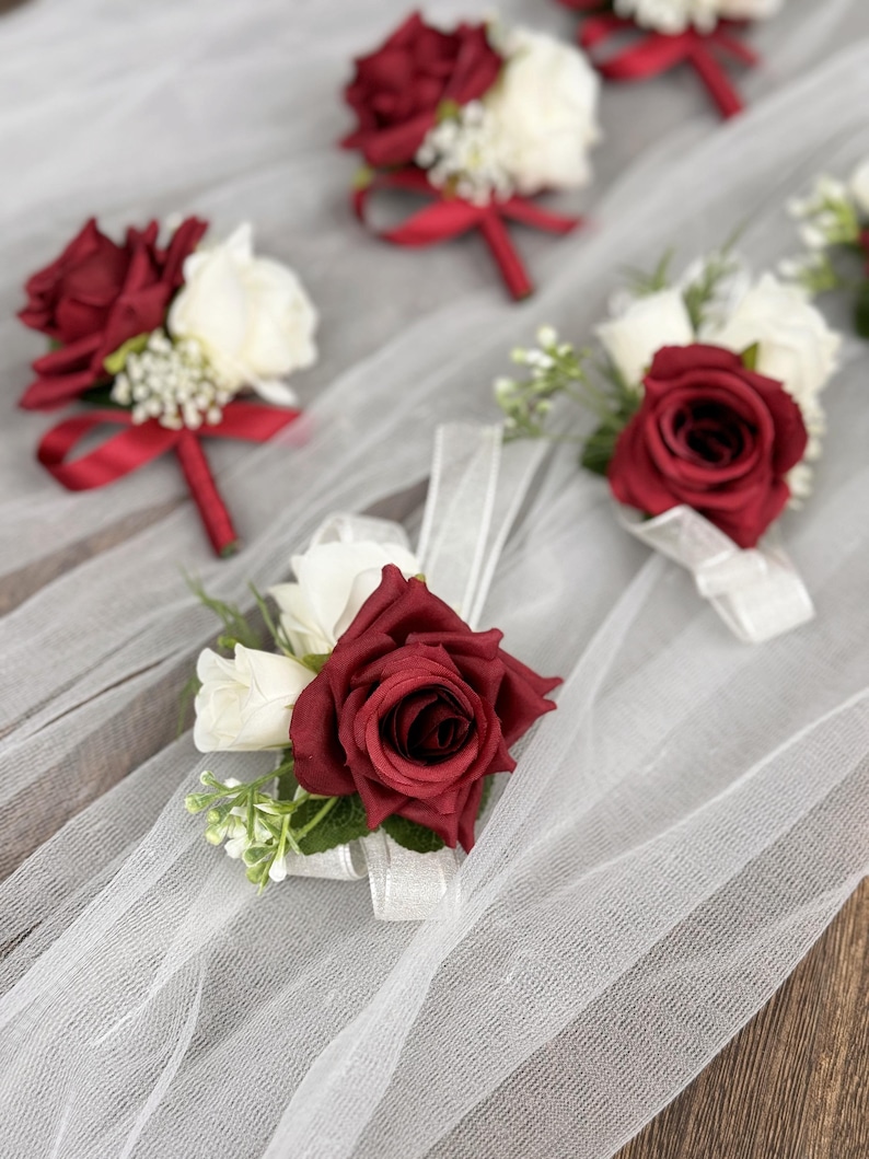 May include: Six burgundy and white artificial flower boutonnieres with red ribbon accents. The boutonnieres are arranged on a white tulle fabric background.