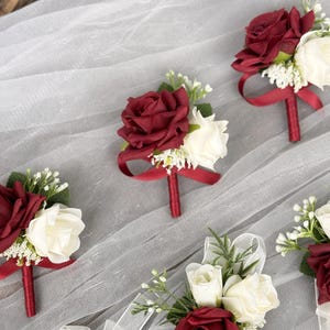 May include: Five burgundy and white rose boutonnieres with burgundy ribbon accents. The boutonnieres are arranged on a white tulle background.