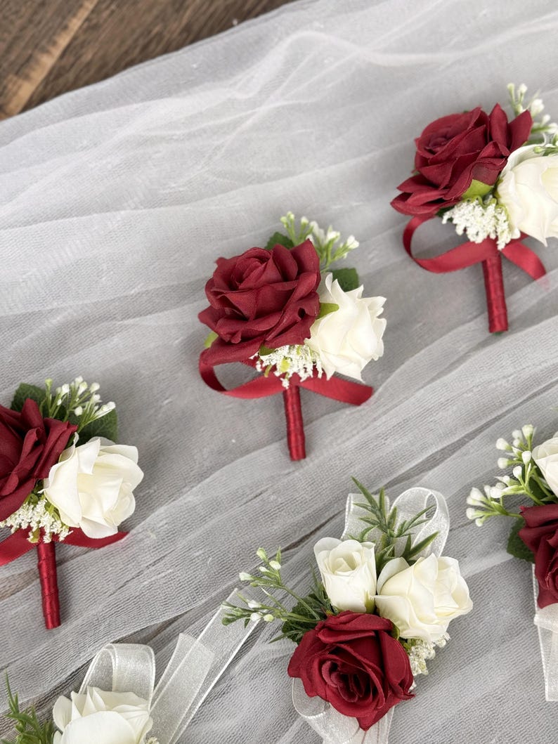 May include: Five burgundy and white rose boutonnieres with burgundy ribbon accents. The boutonnieres are arranged on a white tulle background.