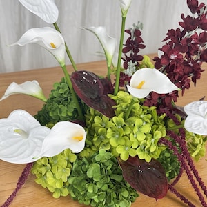 May include: A floral arrangement featuring white calla lilies, white and dark burgundy anthurium flowers, bright green hydrangea clusters, and deep red hanging amaranthus stems, creating a dense, multi-textured botanical display.