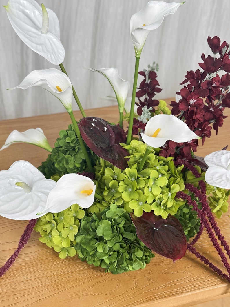May include: A floral arrangement featuring white calla lilies, white and dark burgundy anthurium flowers, bright green hydrangea clusters, and deep red hanging amaranthus stems, creating a dense, multi-textured botanical display.