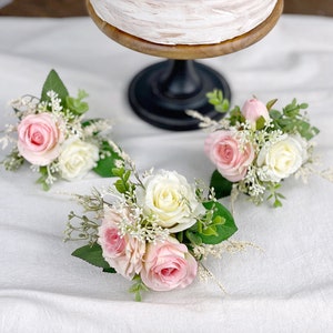 May include: Three small floral arrangements with pink and white roses and greenery. The arrangements are on a white surface.
