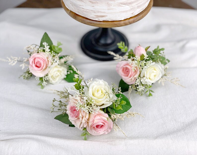May include: Three small floral arrangements with pink and white roses and greenery. The arrangements are on a white surface.