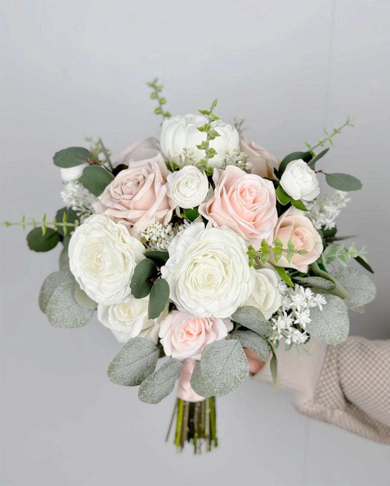 May include: A bridal bouquet featuring white and blush pink roses, white ranunculus, and greenery.