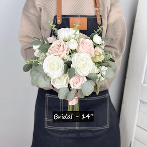 May include: A bridal bouquet featuring white and blush pink roses, greenery, and white baby's breath. The bouquet measures 14 inches in diameter. Bridal - 14"