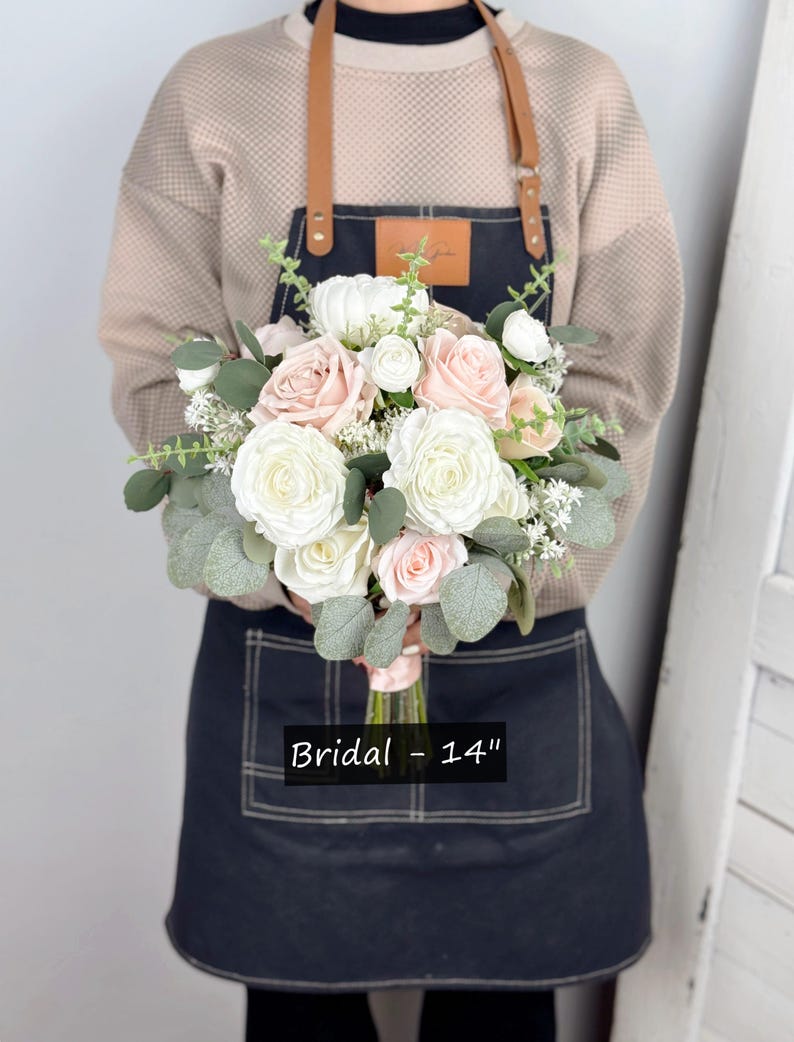 May include: A bridal bouquet featuring white and blush pink roses, greenery, and white baby's breath. The bouquet measures 14 inches in diameter. Bridal - 14"